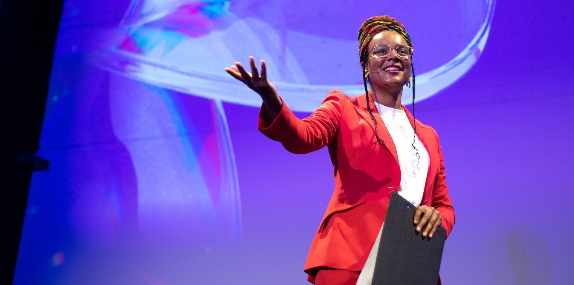 Speaker in a red suit and headscarf gestures with a raised hand while presenting on stage with a headset microphone
